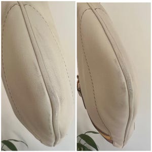 May include: Close-up of a cream-colored leather handbag with brown stitching. The bag has a curved shape and a smooth, slightly textured surface.