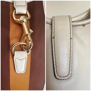 May include: Close-up of a white leather handbag with a gold-toned clasp and brown and orange striped fabric. The bag has a white leather strap with stitching.