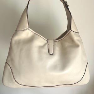 May include: A white leather handbag with a single strap and a gold buckle. The bag has a simple, classic design.