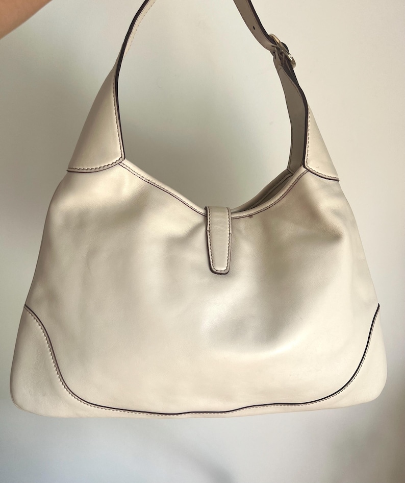 May include: A white leather handbag with a single strap and a gold buckle. The bag has a simple, classic design.
