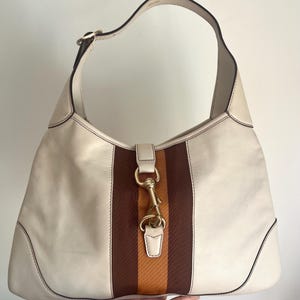 May include: A white leather hobo bag with brown and orange striped fabric accents. The bag has a gold-toned clasp and a single top handle.