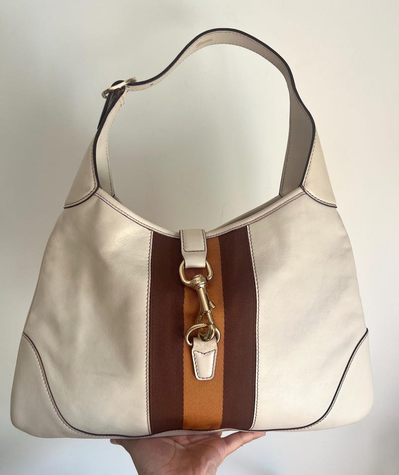 May include: A white leather hobo bag with brown and orange striped fabric accents. The bag has a gold-toned clasp and a single top handle.