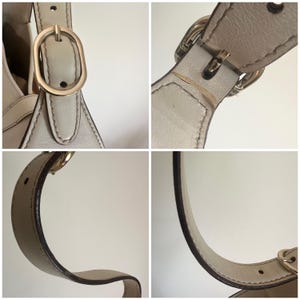May include: A close-up of a light beige leather handbag with a gold buckle and strap. The bag has a simple, elegant design.