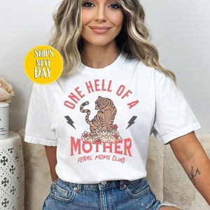 May include: White t-shirt with a graphic of a tiger and the text "One Hell of a Mother Feral Moms Club".