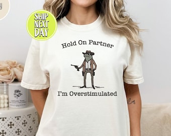 Puede incluir: Una camiseta blanca con un dibujo de una rana de dibujos animados que lleva un sombrero de vaquero y sostiene una pistola. El texto "Hold On Partner I'm Overstimulated" está impreso en la camiseta.