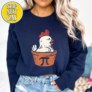 May include: Navy blue sweatshirt featuring a cartoon chicken in a pie crust with the pi symbol. The chicken has a red comb and beak. The sweatshirt has the text "NAVY" on the bottom right and "SHIP NEXT DAY" on the top left.