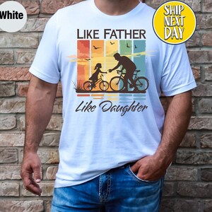May include: White t-shirt with a retro-style graphic of a father and daughter riding bicycles. The graphic features a sunset background with colorful stripes and the text "Like Father Like Daughter".