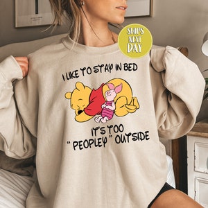 Puede incluir: Una sudadera beige con un gráfico de Winnie de Pooh y Piglet. El texto en la sudadera dice "Me gusta quedarme en la cama. Es demasiado "peopley" afuera."