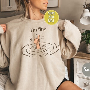 Może przedstawiać: Beżowa bluza z nadrukiem przedstawiającym rękę pokazującą kciuk w górę, podczas gdy jest zanurzona w wodzie. Nad nadrukiem znajduje się napis "I'm fine".