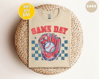 Può includere: Una maglietta beige con il testo "GAME DAY" in lettere blu e rosse. Un guanto da baseball rosso con una palla da baseball bianca è al centro della maglietta. Il guanto è su uno sfondo a scacchi blu e bianchi.
