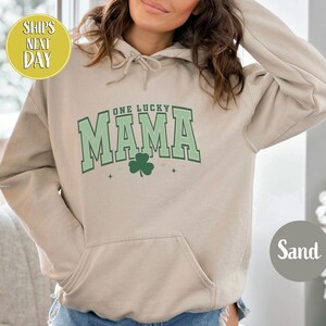 May include: A sand-colored hoodie with a green shamrock and the text "One Lucky Mama" in a green and white varsity style font.