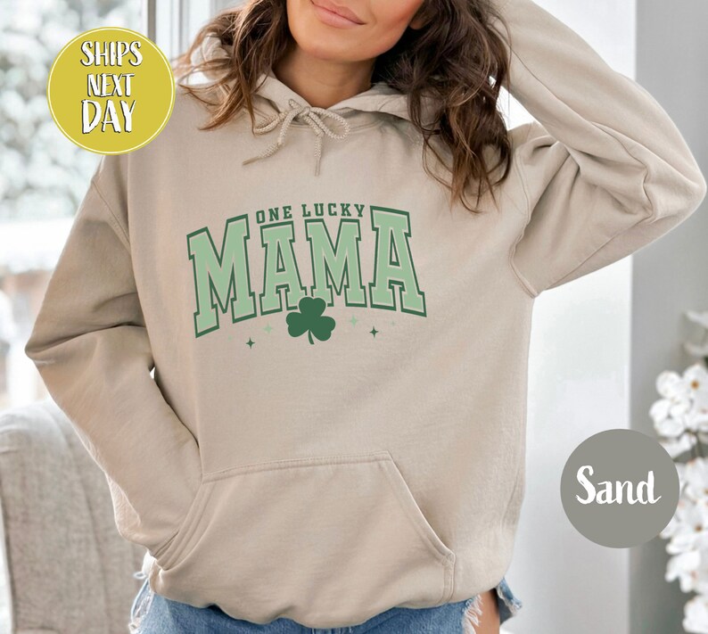 May include: A sand-colored hoodie with a green shamrock and the text "One Lucky Mama" in a green and white varsity style font.