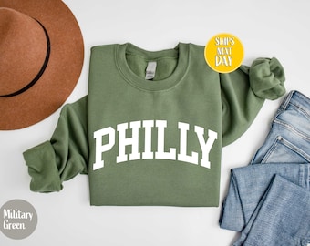 Puede incluir: Una sudadera de cuello redondo en color verde militar con la palabra PHILLY en grandes letras blancas sobre el pecho. La prenda se muestra junto a un sombrero de fieltro marrón y unos vaqueros azules, con una insignia circular amarilla.