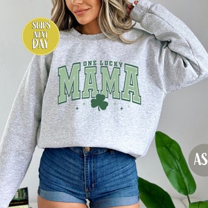 May include: A gray sweatshirt with the text "One Lucky Mama" in green and white lettering. A green shamrock is centered below the text. The sweatshirt is being modeled by a person wearing denim shorts.