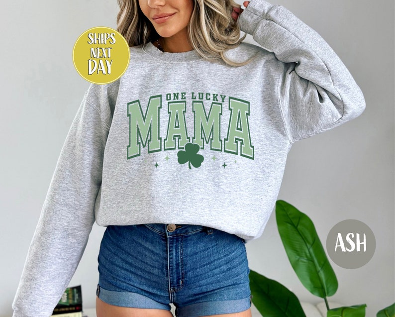May include: A gray sweatshirt with the text "One Lucky Mama" in green and white lettering. A green shamrock is centered below the text. The sweatshirt is being modeled by a person wearing denim shorts.