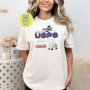 May include: Ivory-colored t-shirt featuring a graphic design with the text "PROTECT THE USPS" in red and blue, along with an illustration of a mail carrier and a mail truck. The phrase "KEEP PUBLIC SERVICES PUBLIC!" is also included.