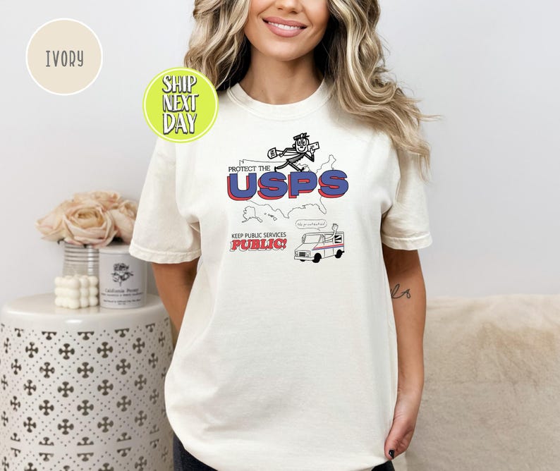May include: Ivory-colored t-shirt featuring a graphic design with the text "PROTECT THE USPS" in red and blue, along with an illustration of a mail carrier and a mail truck. The phrase "KEEP PUBLIC SERVICES PUBLIC!" is also included.