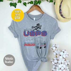 May include: Athletic heather gray t-shirt with a graphic that says "PROTECT THE USPS" in red and blue. The shirt features a cartoon postal worker and a mail truck. The text "KEEP PUBLIC SERVICES PUBLIC!" is also included.