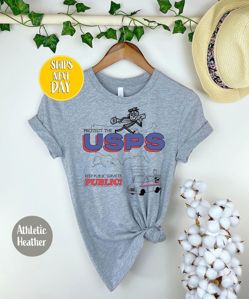 May include: Athletic heather gray t-shirt with a graphic that says "PROTECT THE USPS" in red and blue. The shirt features a cartoon postal worker and a mail truck. The text "KEEP PUBLIC SERVICES PUBLIC!" is also included.