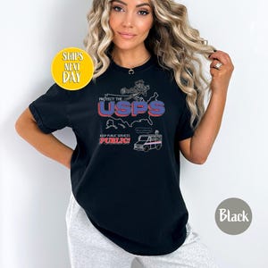 May include: Black t-shirt with a graphic design that reads "Protect the USPS" in red and blue. The design includes a map, a postal worker, and a mail truck. The text "Keep Public Services Public!" is also included.