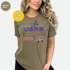 May include: Heather olive-colored t-shirt with a graphic that says "Protect the USPS" in red and blue. The shirt also features a cartoon of a mail carrier and a mail truck. The text "Keep Public Services Public!" is also on the shirt.