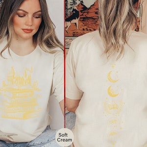 May include: Two women wearing soft cream coloured t-shirts with different graphic designs. The shirt on the left features a yellow illustration of a castle and stacked books. The shirt on the right features a yellow illustration of a crescent moon and stars.