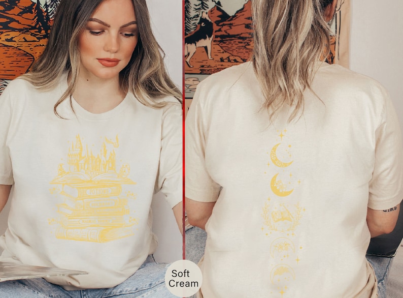 May include: Two women wearing soft cream coloured t-shirts with different graphic designs. The shirt on the left features a yellow illustration of a castle and stacked books. The shirt on the right features a yellow illustration of a crescent moon and stars.