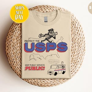 May include: A sand-colored t-shirt with a graphic supporting the USPS. The design features a running cartoon figure over the text "PROTECT THE USPS" and a map of the United States. Below, it reads "KEEP PUBLIC SERVICES PUBLIC!"