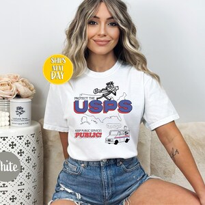 May include: White t-shirt with a graphic that says "Protect the USPS" in red and blue, with a cartoon character and a postal truck. The shirt also has the text "Keep Public Services Public!"