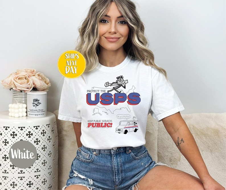 May include: White t-shirt with a graphic that says "Protect the USPS" in red and blue, with a cartoon character and a postal truck. The shirt also has the text "Keep Public Services Public!"