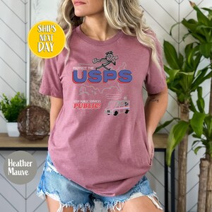 May include: Heather mauve t-shirt with a graphic that says "PROTECT THE USPS" with a cartoon image of a mail carrier and a mail truck. The text "KEEP PUBLIC SERVICES PUBLIC!" is also on the shirt.