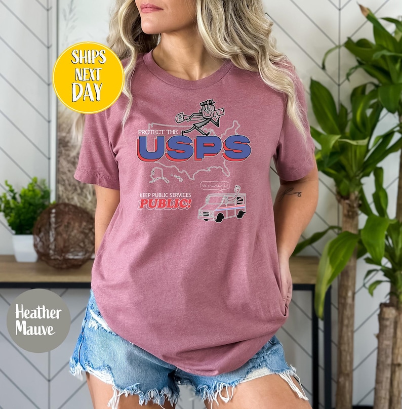 May include: Heather mauve t-shirt with a graphic that says "PROTECT THE USPS" with a cartoon image of a mail carrier and a mail truck. The text "KEEP PUBLIC SERVICES PUBLIC!" is also on the shirt.