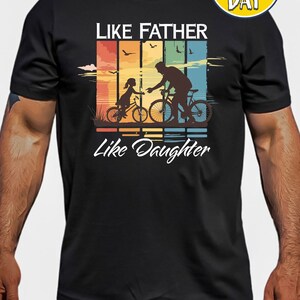 May include: Black t-shirt with a retro-style graphic of a father and daughter riding bicycles. The graphic includes the text "Like Father Like Daughter".