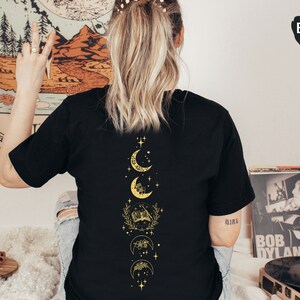 May include: Black t-shirt with a gold moon and stars design on the back. The design features a crescent moon, a full moon, and a half moon with floral accents.