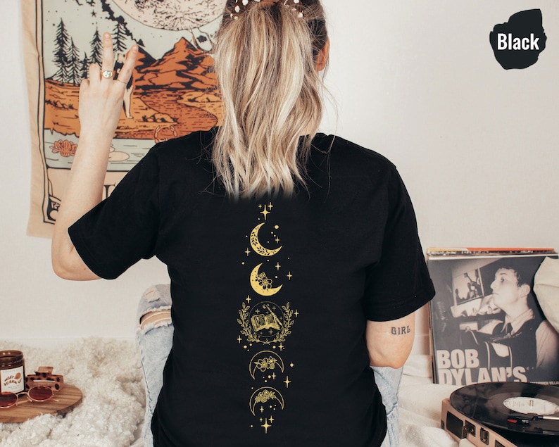 May include: Black t-shirt with a gold moon and stars design on the back. The design features a crescent moon, a full moon, and a half moon with floral accents.