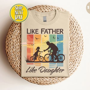 May include: A beige t-shirt with a retro-style graphic of a father and daughter riding bicycles. The graphic features a sunset background with a rainbow and the text "Like Father Like Daughter".