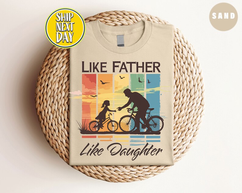 May include: A beige t-shirt with a retro-style graphic of a father and daughter riding bicycles. The graphic features a sunset background with a rainbow and the text "Like Father Like Daughter".