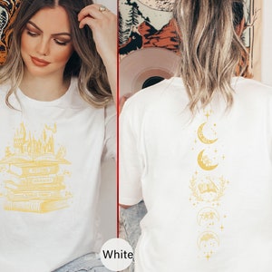 May include: Two white t-shirts with different designs. The first shirt features a stack of books with a castle on top. The second shirt features a vertical design of moons and stars.