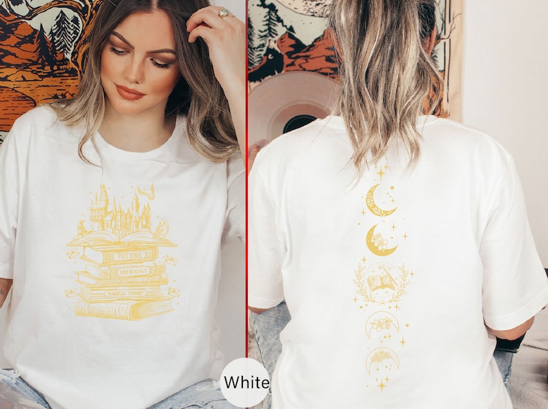 May include: Two white t-shirts with different designs. The first shirt features a stack of books with a castle on top. The second shirt features a vertical design of moons and stars.