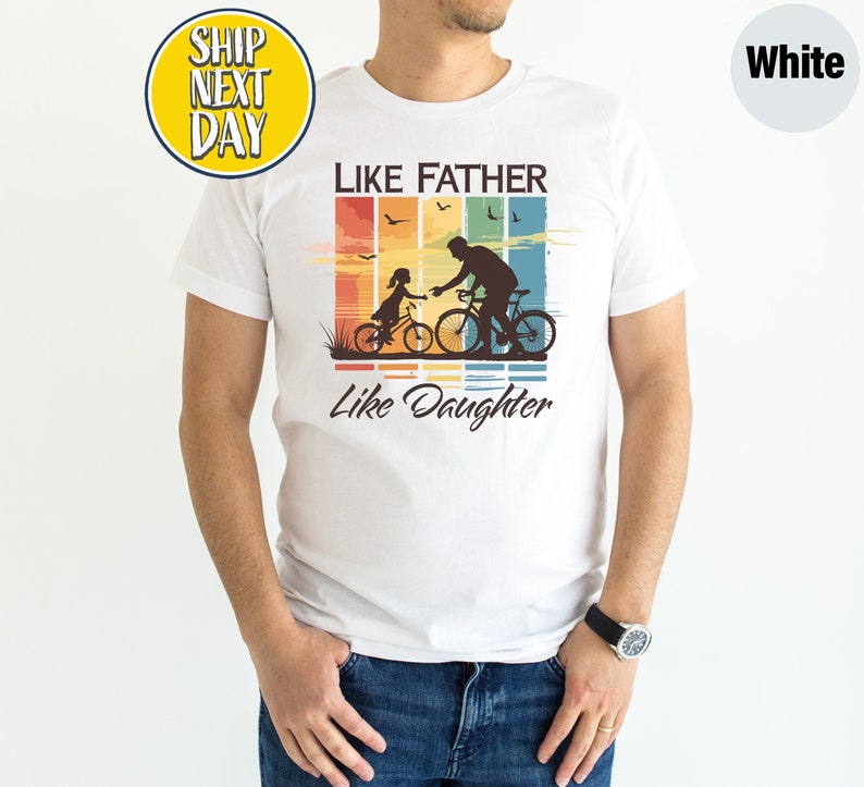 May include: White t-shirt with a vintage-style graphic of a father and daughter riding bicycles. The graphic has a rainbow color scheme and the text "Like Father Like Daughter".