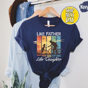 May include: Navy blue t-shirt with a vintage-style graphic of a father and daughter riding bicycles. The graphic has a rainbow color scheme and the text "Like Father Like Daughter".