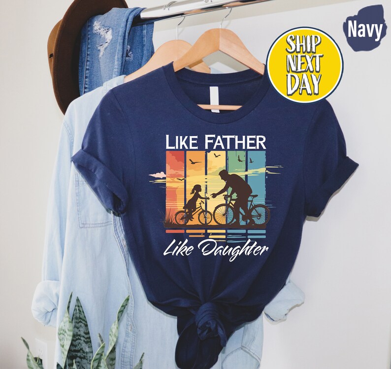 May include: Navy blue t-shirt with a vintage-style graphic of a father and daughter riding bicycles. The graphic has a rainbow color scheme and the text "Like Father Like Daughter".