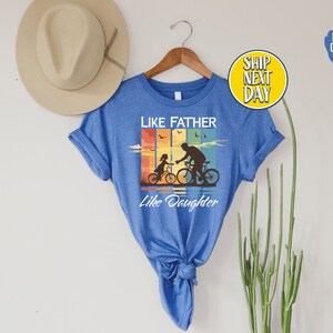 May include: A blue t-shirt with a vintage-style graphic of a father and daughter riding bicycles. The text "Like Father Like Daughter" is printed below the graphic.