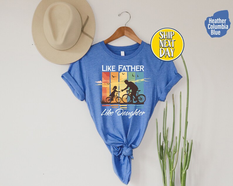 May include: A blue t-shirt with a vintage-style graphic of a father and daughter riding bicycles. The text "Like Father Like Daughter" is printed below the graphic.