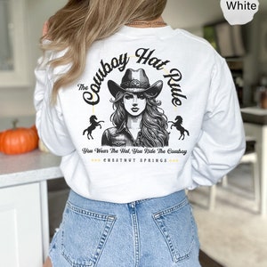 May include: White long-sleeve t-shirt with a graphic of a woman wearing a cowboy hat and riding a horse. The text "The Cowboy Hat Rule" is written in a circle around the woman. The text "You Wear The Hat, You Ride The Cowboy" is written below the graphic. The text "CHESTNUT SPRINGS" is written below the text.