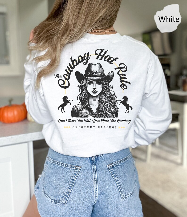 May include: White long-sleeve t-shirt with a graphic of a woman wearing a cowboy hat and riding a horse. The text "The Cowboy Hat Rule" is written in a circle around the woman. The text "You Wear The Hat, You Ride The Cowboy" is written below the graphic. The text "CHESTNUT SPRINGS" is written below the text.