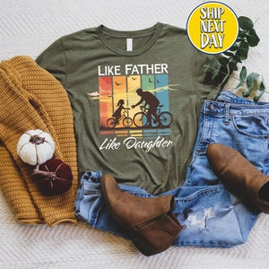 May include: Green t-shirt with a vintage-style graphic of a father and daughter riding bikes. The graphic has a rainbow color scheme and the text "Like Father Like Daughter".