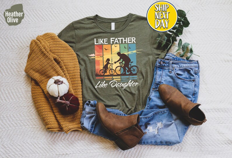 May include: Green t-shirt with a vintage-style graphic of a father and daughter riding bikes. The graphic has a rainbow color scheme and the text "Like Father Like Daughter".