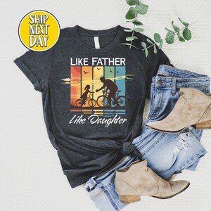 May include: Dark heather grey t-shirt with a vintage-style graphic of a father and daughter riding bicycles. The graphic has a rainbow color scheme and the text "Like Father Like Daughter".