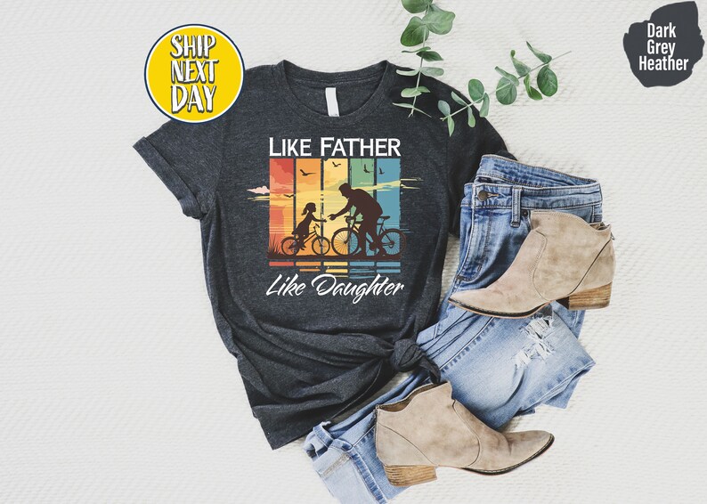 May include: Dark heather grey t-shirt with a vintage-style graphic of a father and daughter riding bicycles. The graphic has a rainbow color scheme and the text "Like Father Like Daughter".
